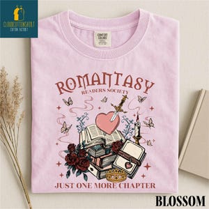 May include: A light pink t-shirt with the words "ROMANTASY READERS SOCIETY" and "JUST ONE MORE CHAPTER". The graphic includes a heart pierced by a sword, books, roses, butterflies, and a crown.