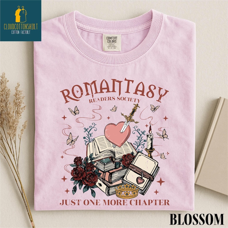 May include: A light pink t-shirt with the words "ROMANTASY READERS SOCIETY" and "JUST ONE MORE CHAPTER". The graphic includes a heart pierced by a sword, books, roses, butterflies, and a crown.