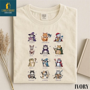 May include: Ivory-colored t-shirt featuring a whimsical design of twelve cats dressed as fantasy characters, including a wizard, barbarian, and bard. The shirt has the text "CLOUDCOTTONSHIRT COTTON FACTORY" on the upper left.