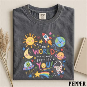 May include: A charcoal gray t-shirt featuring a colorful graphic print of space-themed illustrations including a sun, moon, earth, rocket, planet, alien in a saucer, and astronaut, with the text The WORLD needs more people like YOU!