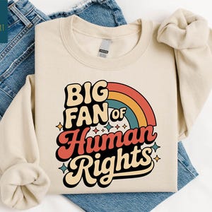 May include: Sand-colored sweatshirt with the text "BIG FAN of Human Rights" in a retro font, featuring a rainbow design. The sweatshirt is laid on top of blue denim jeans.