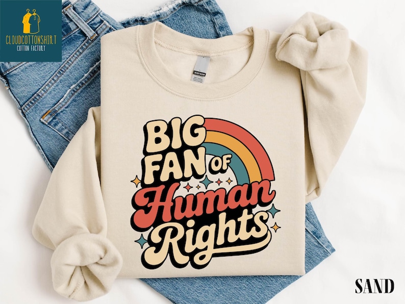May include: Sand-colored sweatshirt with the text "BIG FAN of Human Rights" in a retro font, featuring a rainbow design. The sweatshirt is laid on top of blue denim jeans.