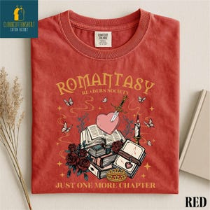 May include: Red t-shirt with the text "ROMANTASY READERS SOCIETY" and "JUST ONE MORE CHAPTER". The graphic includes a heart pierced by a sword, books, butterflies, roses, a crown, and a candle.