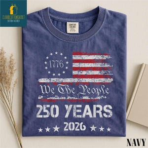 May include: Navy blue t-shirt featuring a distressed American flag design with red and white stripes and stars. The shirt includes the text "1776", "We The People", "250 YEARS", and "2026". The shirt is made of a soft material.