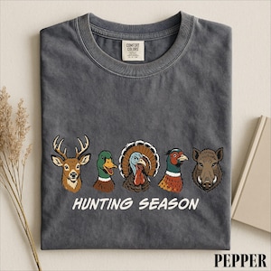 A gray t-shirt with a graphic design featuring four different animal heads, including a deer, a turkey, a boar, and a duck, along with the text "HUNTING SEASON".