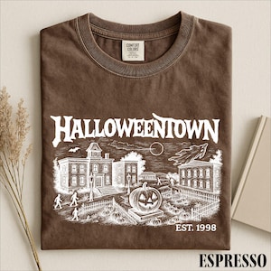 A brown t-shirt with a Halloween-themed graphic design, including a spooky town scene, a jack-o-lantern, and a cemetery.