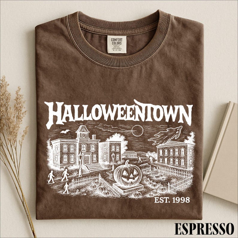 A brown t-shirt with a Halloween-themed graphic design, including a spooky town scene, a jack-o-lantern, and a cemetery.