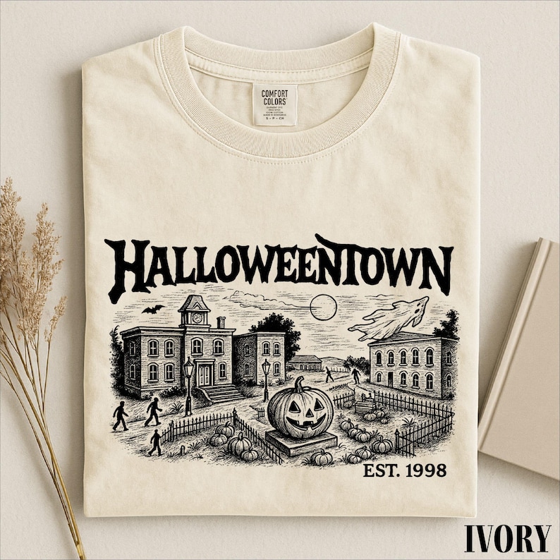 A t-shirt with a Halloween-themed illustration, including a pumpkin patch, buildings, and a ghost, along with the text "HALLOWEENOWN" and "EST. 1998".