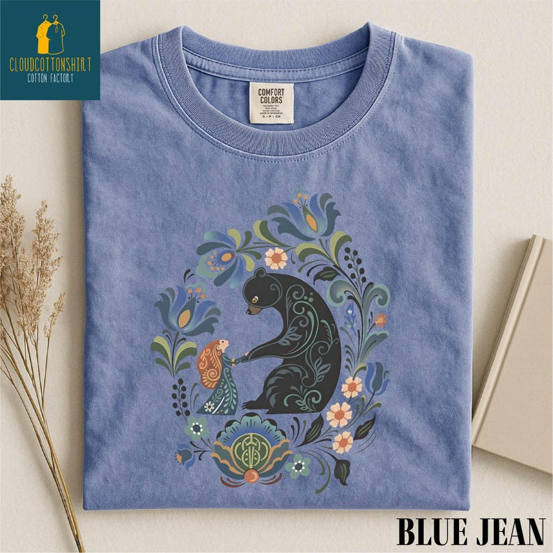 A blue t-shirt with a colorful floral and bear design printed on it, along with some dried flowers and a book.