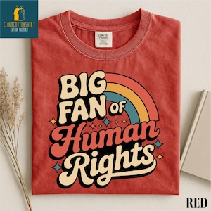 May include: Red t-shirt with the text "BIG FAN of Human Rights" in a retro font, with a rainbow graphic. The shirt has a crew neck and short sleeves. The brand logo is in the upper left corner.