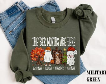Puede incluir: Una sudadera de cuello redondo color verde militar con el texto blanco THE 'BER MONTHS ARE HERE sobre ilustraciones de un árbol otoñal, un fantasma, un pavo, un árbol de Navidad y Santa Claus, con los meses escritos debajo.