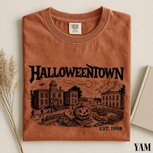 a t-shirt with a Halloween-themed illustration, including a pumpkin patch, buildings, and a full moon.