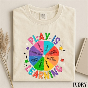 May include: An ivory t-shirt featuring a colorful circular graphic with the text "PLAY IS LEARNING" and segments labeled "MOTOR SKILLS," "SHARING," "EXECUTIVE FUNCTION," "SELF REGULATION," "CREATIVITY," "CURIOSITY," "PROBLEM SOLVING," and "COMMUNICATION."
