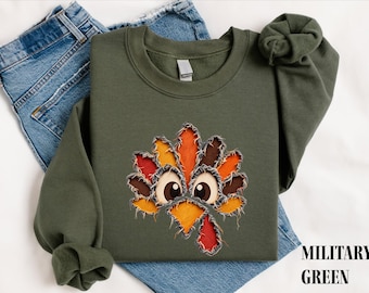 Puede incluir: Una sudadera de cuello redondo color verde militar con una aplicación de bordes deshilachados que muestra una cara de pavo. El diseño está compuesto por piezas de tela superpuestas en tonos naranja, amarillo, marrón y rojo, con ojos grandes.