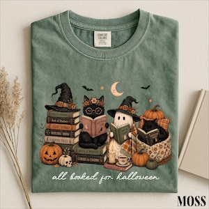 A green t-shirt with a Halloween-themed illustration of a witch, cat, ghost, pumpkin, and books, along with the text "all read for Halloween" and a label that says "COMFORT CLUB".