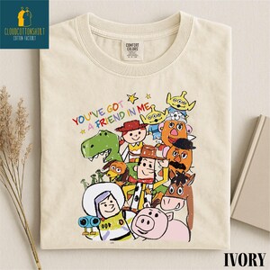 K&ouml;nnte beinhalten: Elfenbeinfarbenes T-Shirt mit einer farbenfrohen Illustration von Zeichentrickfiguren und dem Text "YOU'VE GOT A FRIEND IN ME". Das Shirt hat einen Rundhalsausschnitt und ein Comfort Colors-Etikett.