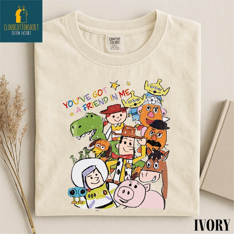 K&ouml;nnte beinhalten: Elfenbeinfarbenes T-Shirt mit einer farbenfrohen Illustration von Zeichentrickfiguren und dem Text "YOU'VE GOT A FRIEND IN ME". Das Shirt hat einen Rundhalsausschnitt und ein Comfort Colors-Etikett.