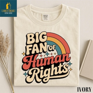 May include: Ivory-colored t-shirt with the text "BIG FAN of Human Rights" in a retro font, featuring a rainbow design. The shirt has a crew neck and is made of a soft material. The shirt also has a tag that says "Comfort Colors".