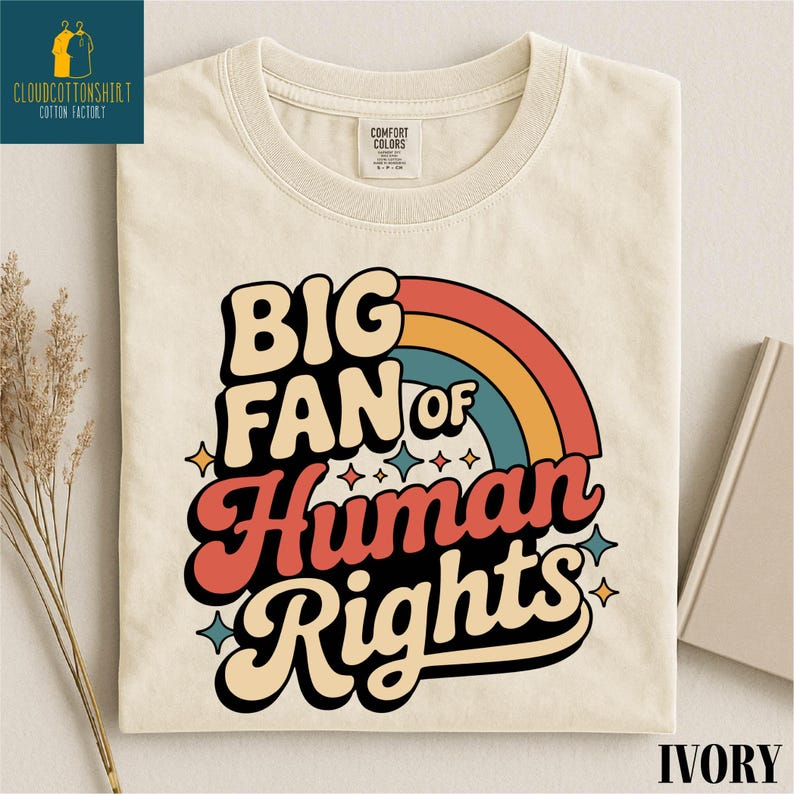 May include: Ivory-colored t-shirt with the text "BIG FAN of Human Rights" in a retro font, featuring a rainbow design. The shirt has a crew neck and is made of a soft material. The shirt also has a tag that says "Comfort Colors".