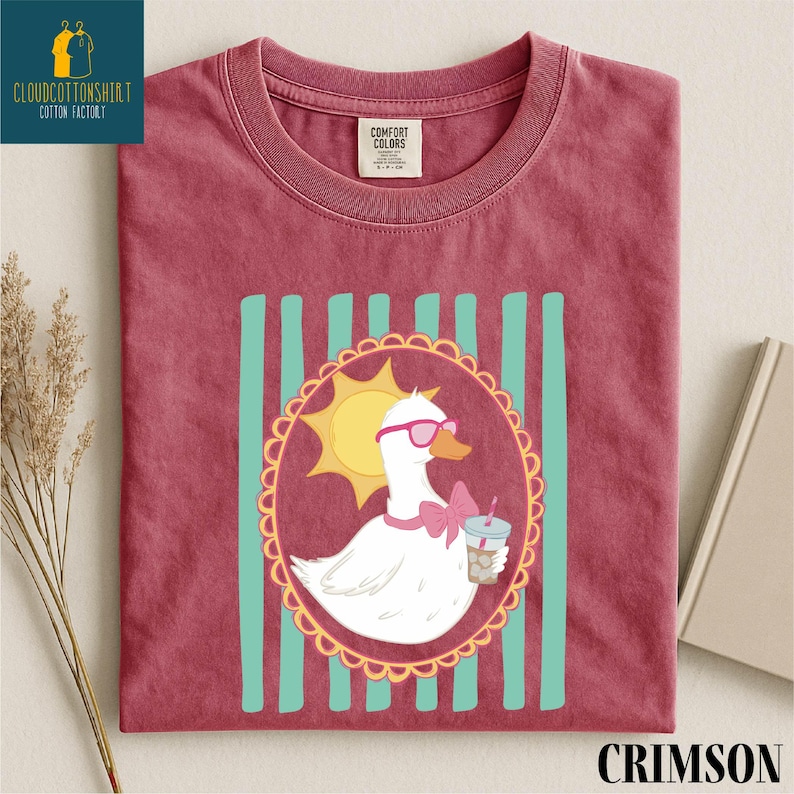 A red t-shirt with a cartoon image of a duck wearing sunglasses and holding a drink, surrounded by a decorative border.