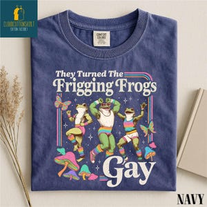 Pu&ograve; includere: T-shirt blu navy con grafica di tre rane danzanti, farfalle e funghi. Il testo recita "They Turned The Frigging Frogs Gay". La maglietta ha un girocollo e un'etichetta Comfort Colors.