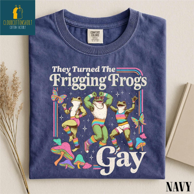 Pu&ograve; includere: T-shirt blu navy con grafica di tre rane danzanti, farfalle e funghi. Il testo recita "They Turned The Frigging Frogs Gay". La maglietta ha un girocollo e un'etichetta Comfort Colors.