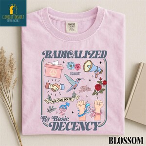May include: A light pink t-shirt with a graphic design featuring the words "RADICALIZED By BASIC DECENCY". The design includes illustrations of a hummingbird, flowers, a megaphone, and other symbols. The shirt is labeled "BLOSSOM".