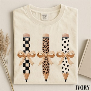 a white t-shirt with a graphic design of three pencils with different patterns and bows.