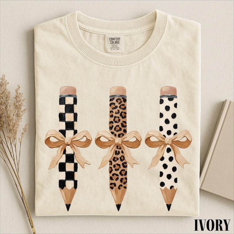 a white t-shirt with a graphic design of three pencils with different patterns and bows.