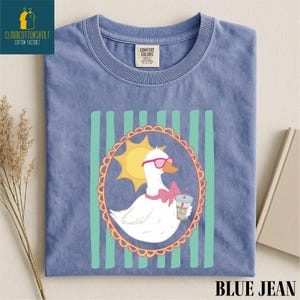 A blue t-shirt with a cartoon image of a duck wearing sunglasses and holding a drink, surrounded by a decorative border.