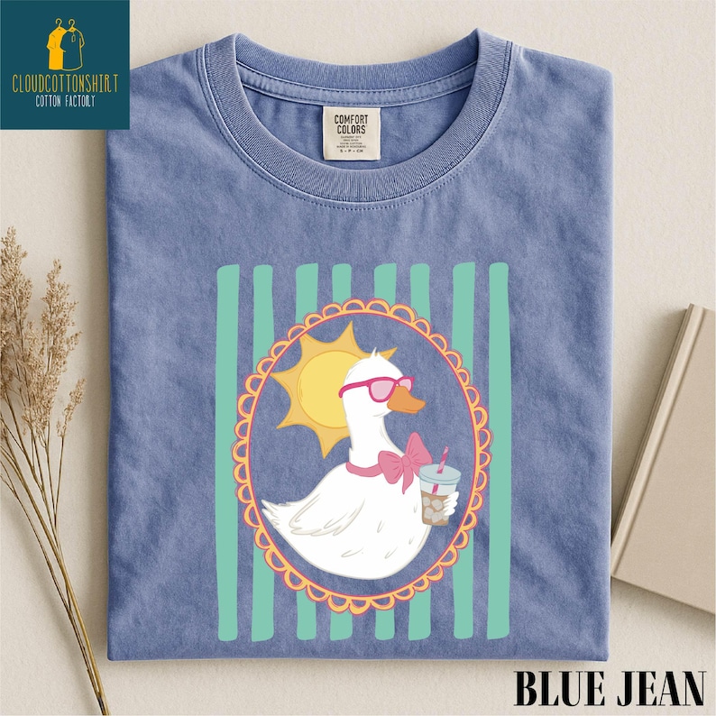 A blue t-shirt with a cartoon image of a duck wearing sunglasses and holding a drink, surrounded by a decorative border.