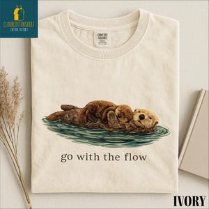 a white t-shirt with a printed illustration of two brown otters swimming together in a body of water. The text below the illustration reads "go with the flow".