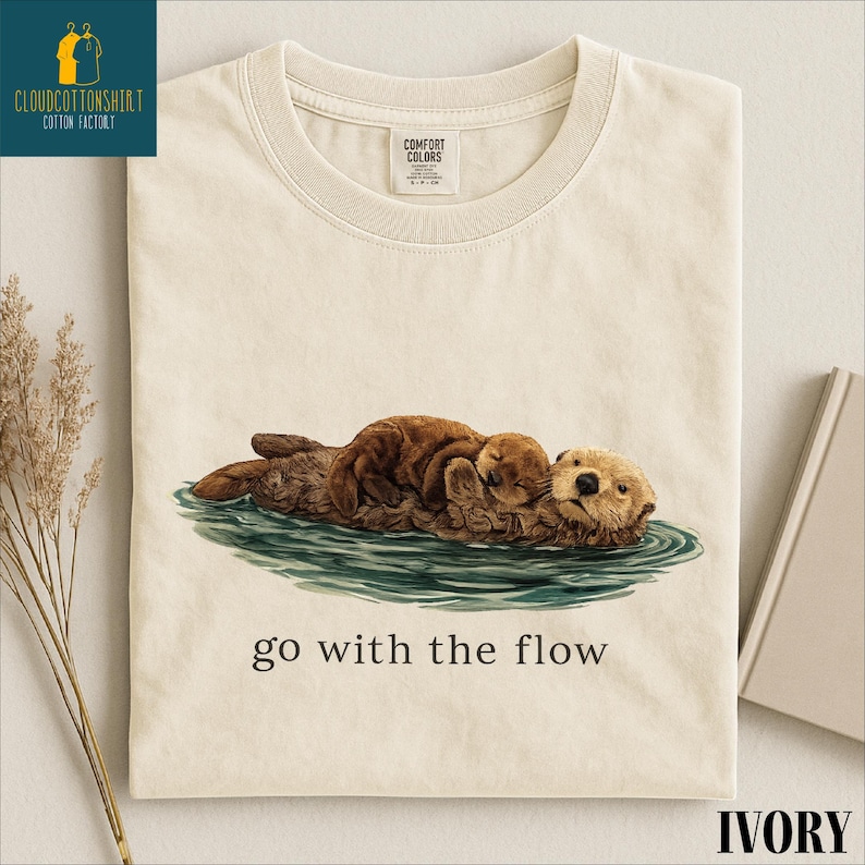 a white t-shirt with a printed illustration of two brown otters swimming together in a body of water. The text below the illustration reads "go with the flow".