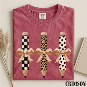a pink t-shirt with three pencils, each with a unique design, and bows tied around them.