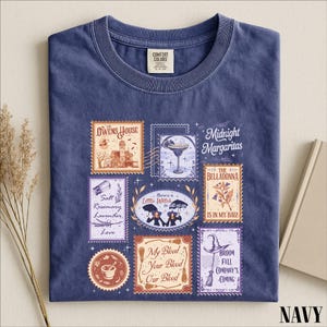 May include: A navy blue t-shirt featuring a graphic print of multiple postage stamp-style illustrations. The designs include a house, a margarita, a mortar and pestle, brooms, and text such as The Owens House, Midnight Margaritas, and My Blood Your Blood Our Blood.