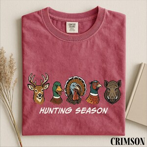 A red t-shirt with a graphic design of five animal heads, including a deer, duck, turkey, boar, and pheasant, along with the text "HUNTING SEASON".