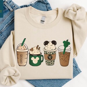 May include: Sand-colored sweatshirt featuring four stylized coffee drinks. Designs include a Starbucks-style cup, a green mug with a Mickey Mouse design, a cup with a leopard print, and a clear cup with a green straw. The word "SAND" is printed at the bottom.