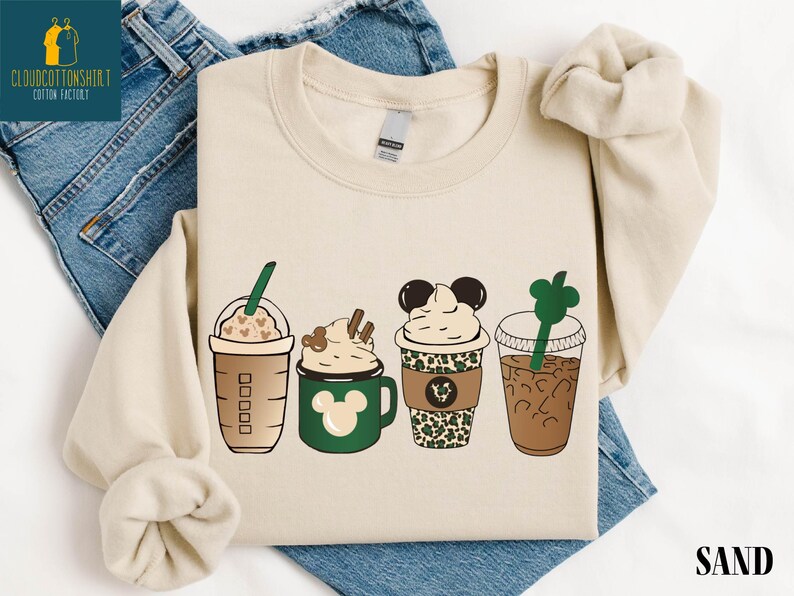 May include: Sand-colored sweatshirt featuring four stylized coffee drinks. Designs include a Starbucks-style cup, a green mug with a Mickey Mouse design, a cup with a leopard print, and a clear cup with a green straw. The word "SAND" is printed at the bottom.
