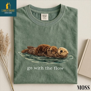 A green t-shirt with an illustration of two otters swimming in a body of water, accompanied by the text "go with the flow" printed on it.