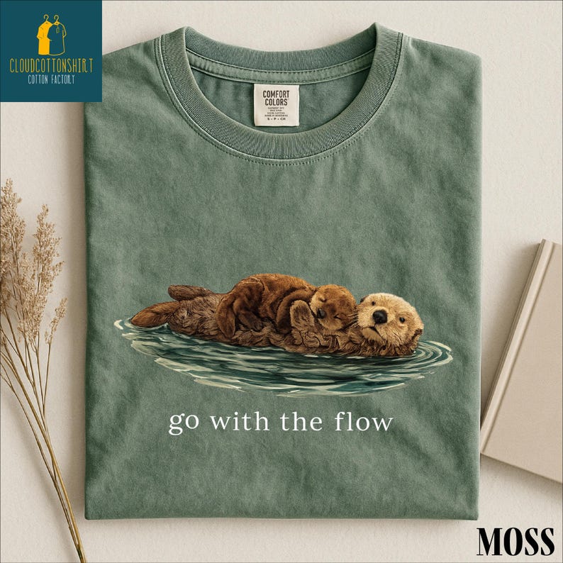 A green t-shirt with an illustration of two otters swimming in a body of water, accompanied by the text "go with the flow" printed on it.