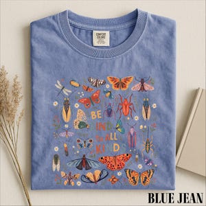 A blue t-shirt with a colorful graphic design featuring various insects and butterflies, along with the text "Be kind to all kids".