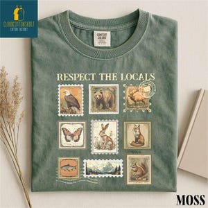 May include: Moss green t-shirt with the text "RESPECT THE LOCALS" and vintage-style postage stamp graphics of wildlife, including an eagle, bear, deer, butterfly, rabbit, fox, fish, and squirrel. The shirt has a crew neck.