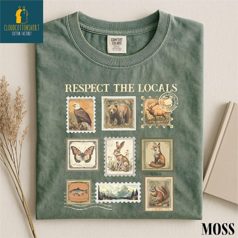 May include: Moss green t-shirt with the text "RESPECT THE LOCALS" and vintage-style postage stamp graphics of wildlife, including an eagle, bear, deer, butterfly, rabbit, fox, fish, and squirrel. The shirt has a crew neck.