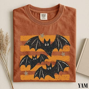 May include: A rust-colored short-sleeve t-shirt featuring a graphic print of three black bats flying over horizontal orange stripes and small gray stars. The shirt has a crew neckline and a Comfort Colors brand label on the inside collar.