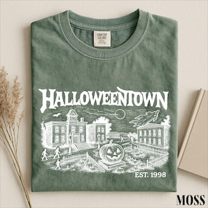 A green t-shirt with a Halloween-themed illustration and text, including the words "Halloweentown" and "EST. 1998", along with a small pumpkin and a bat.