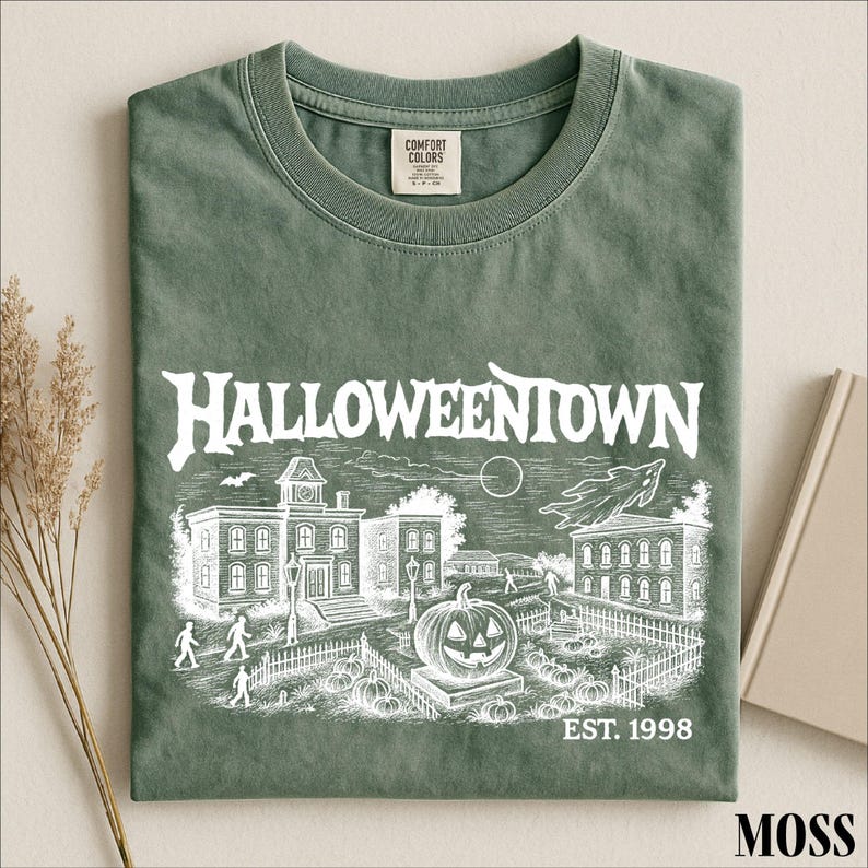 A green t-shirt with a Halloween-themed illustration and text, including the words "Halloweentown" and "EST. 1998", along with a small pumpkin and a bat.