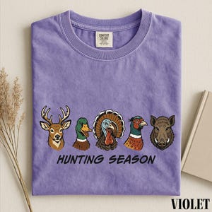 A purple t-shirt with a graphic design of various hunting animals, including a deer, turkey, duck, and boar, along with the text "HUNTING SEASON".