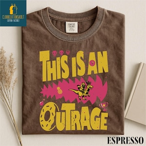 a brown t-shirt with a graphic design that says "THIS IS AN OUTRAGE" in bold yellow and pink letters, along with a dinosaur and other colorful elements.