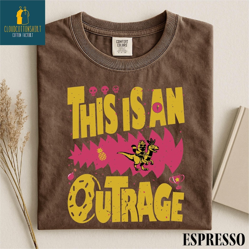 a brown t-shirt with a graphic design that says "THIS IS AN OUTRAGE" in bold yellow and pink letters, along with a dinosaur and other colorful elements.