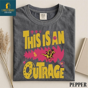 a gray t-shirt with a vibrant graphic design, including a dinosaur, flowers, and the text "This is an Outrage".